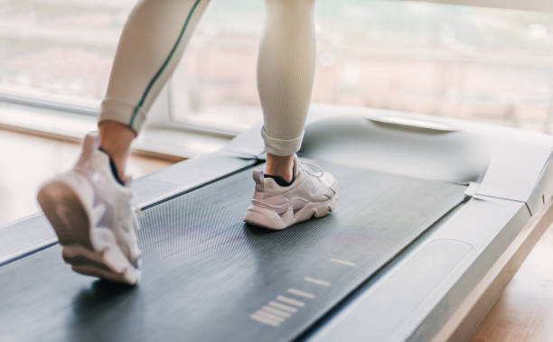 détail flou sélectif des jambes qui courent sur un tapis roulant. une lumière chaude pénètre par la fenêtre en arrière-plan. - treadmill photos et images de collection