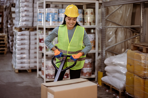 Asian female Worker pulling pallet jack with boxes in a large Warehouse
