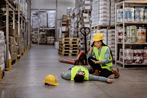 Female supervisor crouching while talking through mobile phone by unconscious male worker lying on floor at warehouse