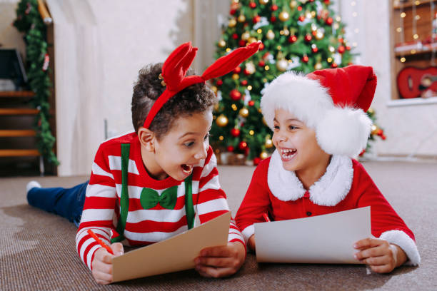 un garçon noir et une fille sont allongés sur le sol près d’un arbre de noël décoré et lisent des lettres pour le père noël - christmas child baby surprise photos et images de collection