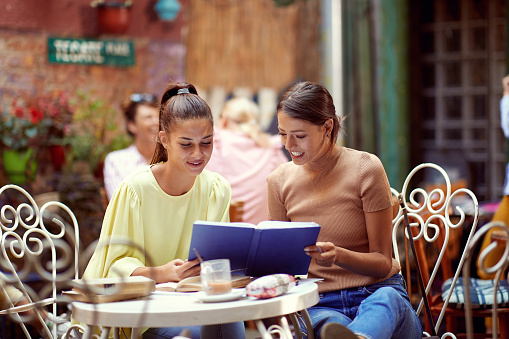 young adult caucasian females reading notebook together, smiling, sitting  in the outdoor cafe
