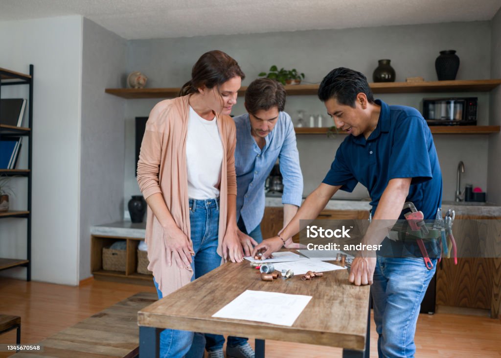 Plumber fixing a pipe and talking to his clients in the kitchen Happy Latin American plumber fixing a pipe and talking to his clients in the kitchen - domestic life concepts Building Contractor Stock Photo