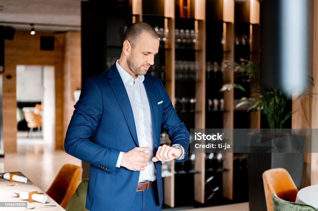 Businessman checking the time in a restaurant Businessman standing and checking the time on his wrist watch in a restaurant. Businessman Stock Photo