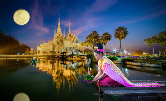 Beautiful Thai woman in Thai traditional costume on the wooden floor and holding floating basket (Krathong in Thai language) for floating in the red lotus lake in Loy Krathong Festival of Thailand.