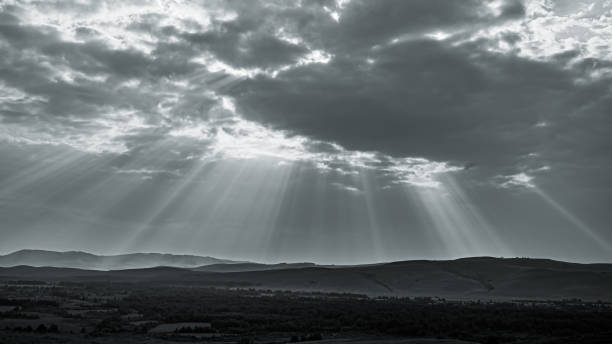 istock Sun rays through cloudy sky. Visible sun light columns above dark mountain landscape in black and white tones 1345686078