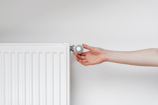 Cropped view of woman hand adjusting thermostat on white heating radiator. Consumption of natural resource, saving energy and warm home concept