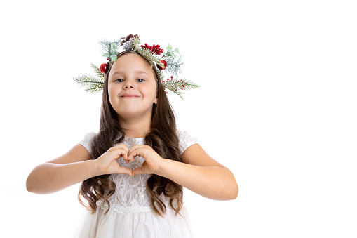 portrait of charming, cheerful girl in Christmas wreath, white dress showing heart sign with hands, isolated on white background with copy space.