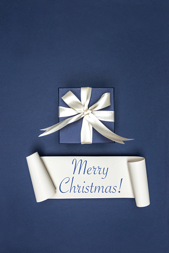 Blue gift box with white ribbon on blue background and white card with Merry Christmas text.