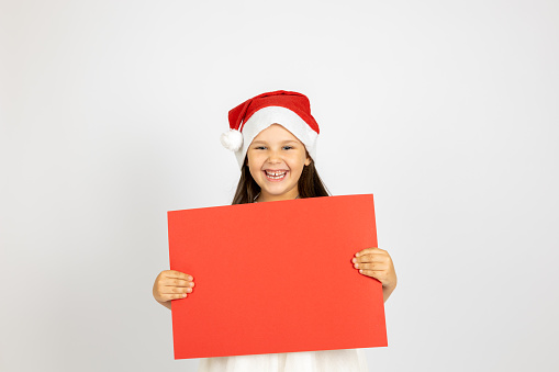 portrait of laughing, Caucasian girl in white dress and santa Claus hat holding red blank poster isolated on white background.