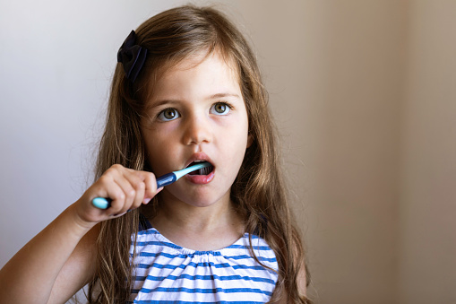 Caucasian toddler girl is brushing her teeth in front of a blank gray wall and is looking up.