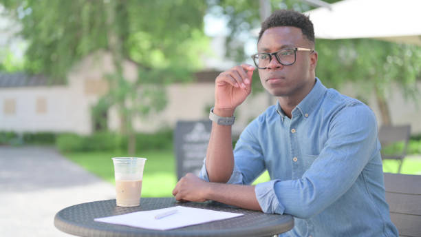 jeune homme africain pensif avec du café et des documents, pensant dans un café en plein air - 24296 photos et images de collection