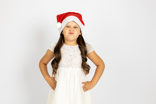 portrait of frowning, dissatisfied girl in Santa Claus hat with hands on waist, isolated on white background, concept of dissatisfaction with New Year gift.