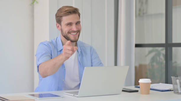istock Creative Man with Laptop Pointing at Camera in Office 1345717217