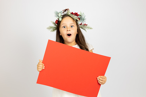 portrait of surprised, shocked girl with mouth open in Christmas wreath holding red blank banner with copy space, isolated on white background.