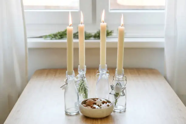 Four candles in bottles is lit for Advent, Christmas cookies and decoration on a table near the window, copy space, selected focus, narrow depth of field