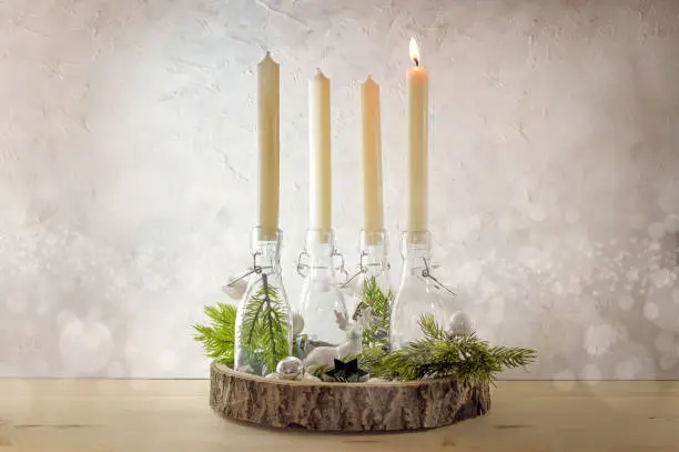 Creative Advent wreath, candles in bottles on a wooden board with fir branches and decoration against a light rustic wall, one is lit at the first Sunday before Christmas, copy space, selected focus