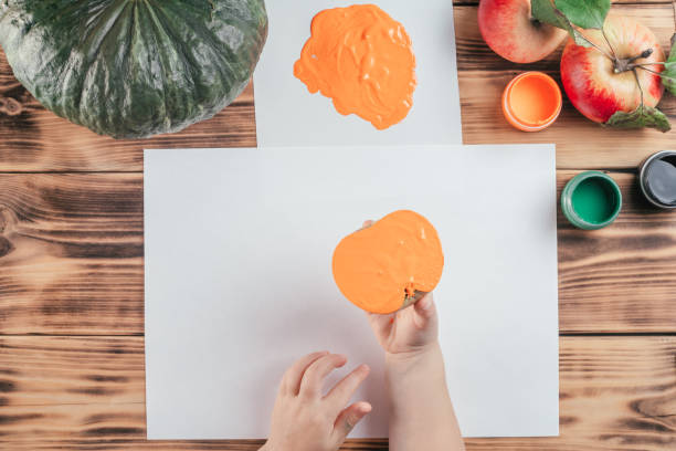 istock Step-by-step Halloween tutorial pumpkin apple prints. Step 7: Child hand holds half of apple painted in orange gouache 1345735316