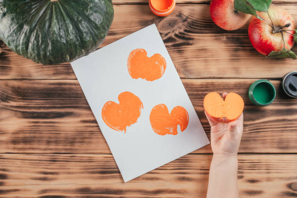 istock Step-by-step Halloween tutorial pumpkin apple prints. Step 10: Child hand holds half of apple painted in orange gouache 1345735326