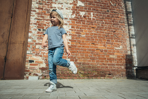 Little cute girl in a blue t-shirt and jeans with a cap is dancing on the background of an old brick wall.