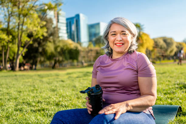 зрелая женщина улыбается и пьет воду после тренировки йоги в городском парке - senior adult outdoors wellbeing sky стоковые фото и изображения