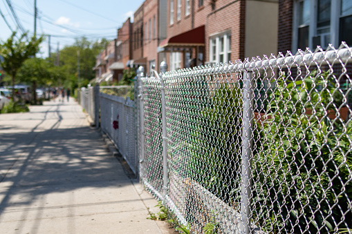 A chain link fence along an empty sidewalk with a row of old brick homes with gardens in Astoria Queens New York