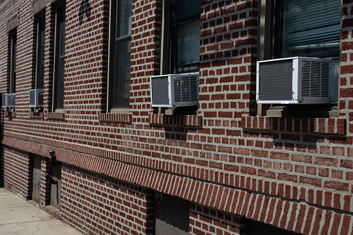 A row of window air conditioners outside a generic old brick apartment building along a sidewalk in Astoria Queens New York