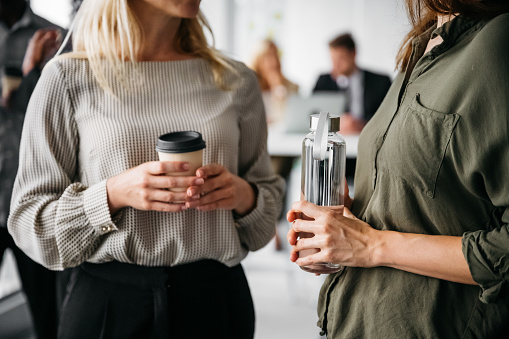 A close-up shot of two smartly dressed business women holding a water bottle and a paper coffee cup. Their faces are not visible, the focus is on their hands. Horizontal daylight indoor photo.