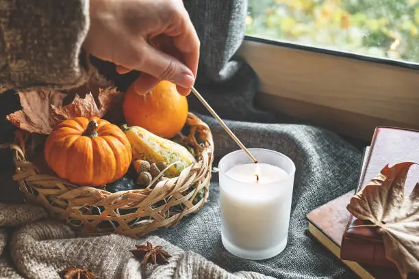 Photo of Hand with burning match lighting a candle on the windowsill with cozy autumn still life with pumpkins, knitted woolen sweater and books. Autumn home decor. Cozy fall mood. Thanksgiving. Halloween.