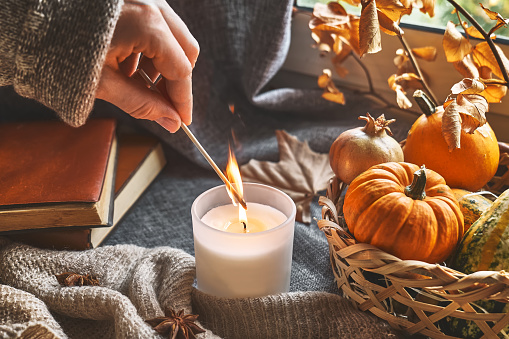 Hand with burning match lighting a candle on the windowsill with cozy autumn still life with pumpkins, knitted woolen sweater and books. Autumn home decor. Cozy fall mood. Thanksgiving. Halloween.