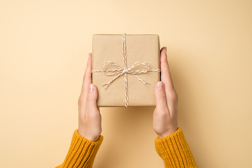 First person top view photo of hands in orange sweater holding craft paper giftbox with twine bow on isolated beige background