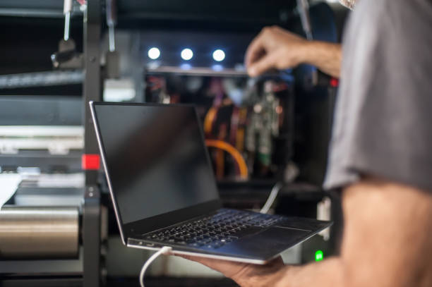 A person is holding a laptop with a blank screen while adjusting components inside a machinery unit that features colorful wires and illuminated lights.
