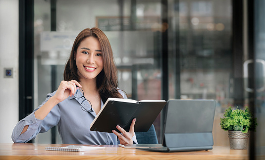 Portrait of beautiful asian woman holding book and glasses, smiling and looking at camera while sitting at office desk.