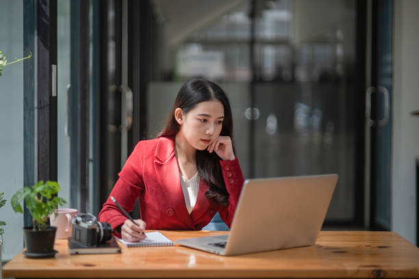 istock Businesswoman writing a note while using laptop at a table in an office. 1345809830