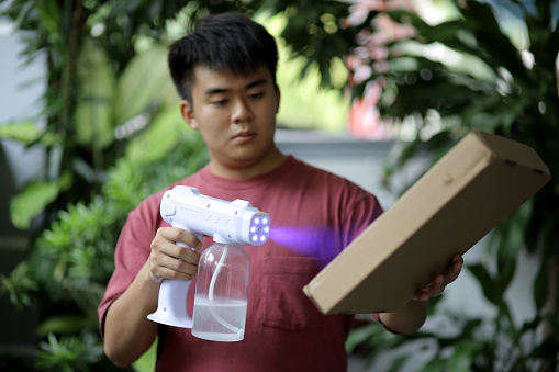 An Asian young man is using disinfectant nano spray on freshly received parcel delivery to him.