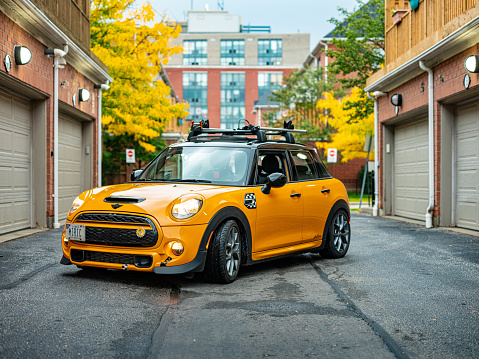 Toronto, Ontario, Canada- October 09, 2021. Volcanic orange colour MINI COOPER  in the city neighbourhood  at West side of Toronto, Canada. MINI is slightly modified and has double bike racks on the roof. This is the third generation model F55 (4 doors), since BMW took over iconic brand of MINI. For the first time, this compact car features engine build and designed by BMW, and packs even more power and torque than previous models since 2002 to present. Original design clues and themes are still present on this brand new model. Mini has been around since 1959 and has been owned and issued by various car manufacturers.