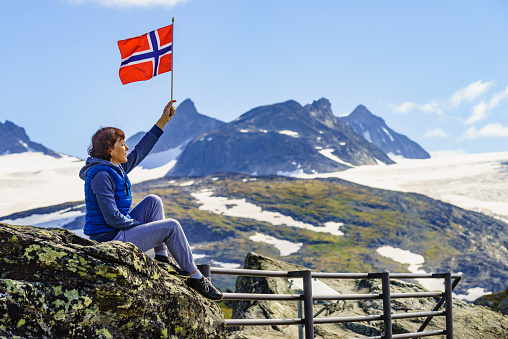 Tourist woman enjoy mountains landscape, holding norwegian flag. National tourist scenic route 55 Sognefjellet, Norway