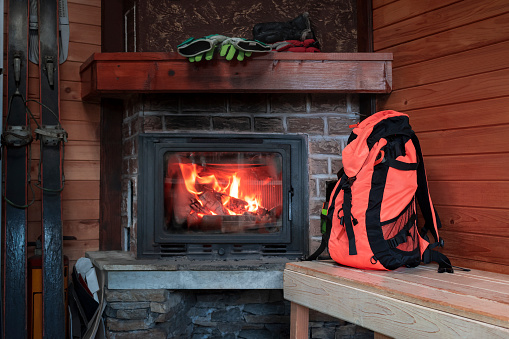 Fireplace in ski forest lodge with tourist backpack and ski. Cozy old fireside in winter hut in mountains at ski resort. Rest and relaxation area after winter sports activity