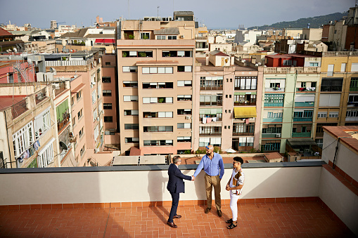 High angle full length view of mature salesman shaking hands with mid adult gay couple as they stand on outdoor balcony with view of residential district.