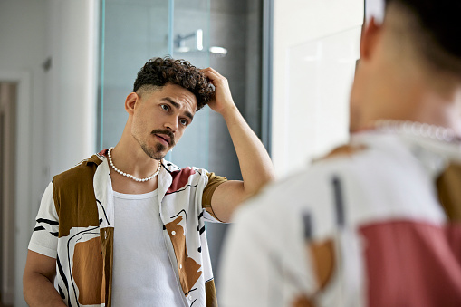 Over the shoulder waist-up view of brunette man in casual clothing standing in modern bathroom and looking at his reflection with hand in hair.