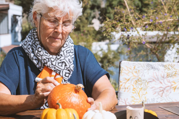 Concentrated senior woman is decorating on a Halloween pumpkin Preparing Halloween decoration. Female person with grey hair is decorating a pumpkin for the celebration. A group of organic fresh vegetables are in front of her. Crafts and hobby. pumpkin decorating stock pictures, royalty-free photos & images