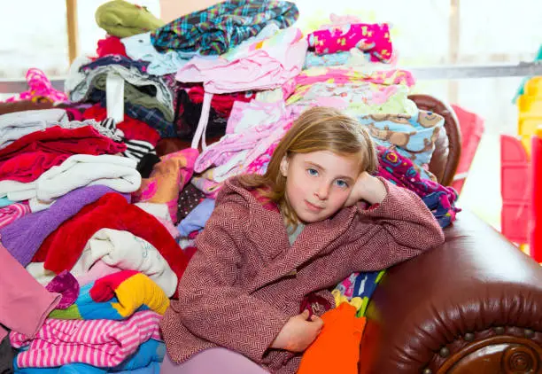 Blond kid girl sitting on a messy clothes sofa before folding laundry