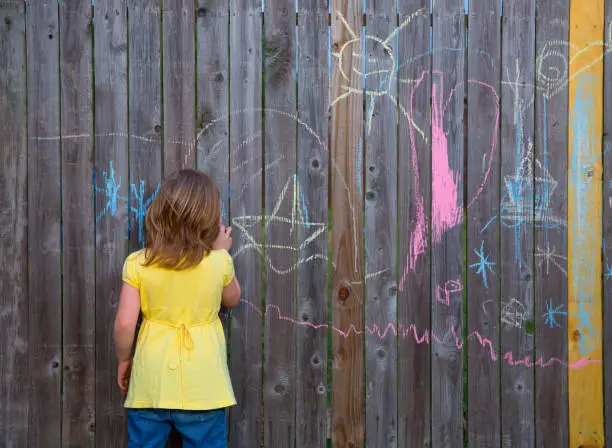 Blond kid girl playing with drawing chalks in the backyard wooden fence rear view