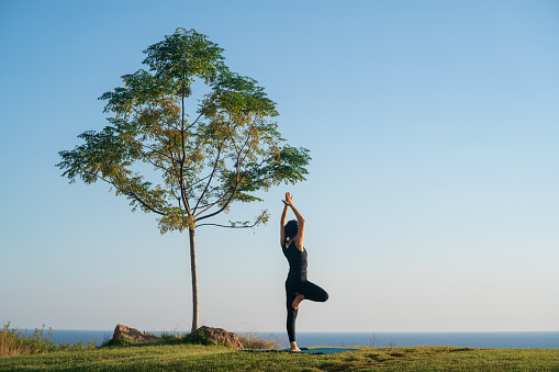 Full length photo of adult woman practicing yoga exercises in outdoor. She is by sea under a tree. Sky is clear and blue. Mediterranean Sea is seen on the background. Model is standing next to tree seen in full length. Shot with a full frame mirrorless camera in outdoor.