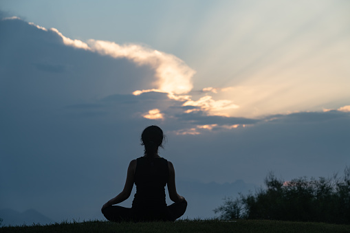 Photo of adult woman meditating during sunset in outdoor. Shot in silhouette with a full frame mirrorless camera. Dominant color is orange due to time of the day.
