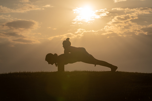 Silhouette photo of adult woman teaching little girl yoga exercises. The background is orange sunset sky. Shot with a full frame mirrorless camera.
