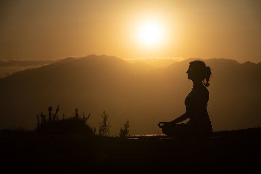 Photo of adult woman meditating during sunset. Shot in silhouette with a full frame mirrorless camera. Dominant color is orange due to time of the day.