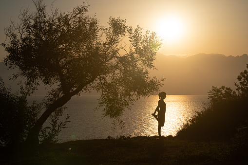 Photo of adult woman doing yoga exercises during sunset. Shot in silhouette with a full frame mirrorless camera. Dominant color is orange due to time of the day.