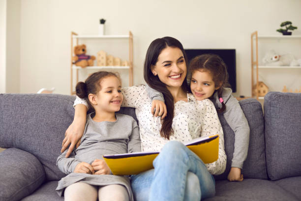 istock Smiling cheerful mother and small daughters sitting on sofa with book, hugging and looking aside 1345852129