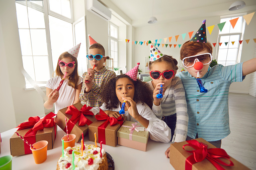 Group of happy diverse children in party hats and funny sunglasses blowing noisemakers and looking at camera while having fun during birthday celebration at home