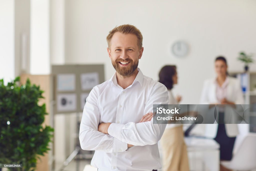 Happy successful businessman or company employee standing in office looking at camera Portrait of happy successful handsome businessman in office workspace. Confident bearded businessman or head of company department standing arms crossed looking at camera and smiling Founder Stock Photo